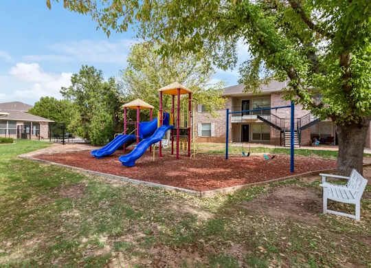 A playground with a blue slide and a yellow structure is surrounded by a grassy area and a tree.