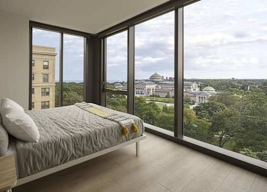 A bedroom with a large bed and a view of a cityscape through the windows.