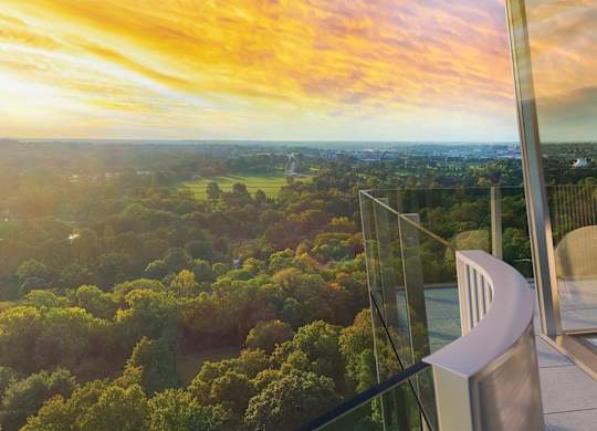 A glass balcony overlooks a forest and a cityscape at sunset.
