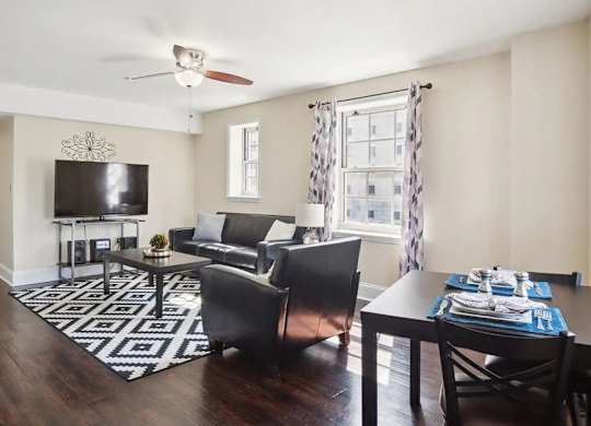 A living room with a black and white rug, a black couch, a black chair, a black coffee table, a flat screen TV, and a window with curtains.