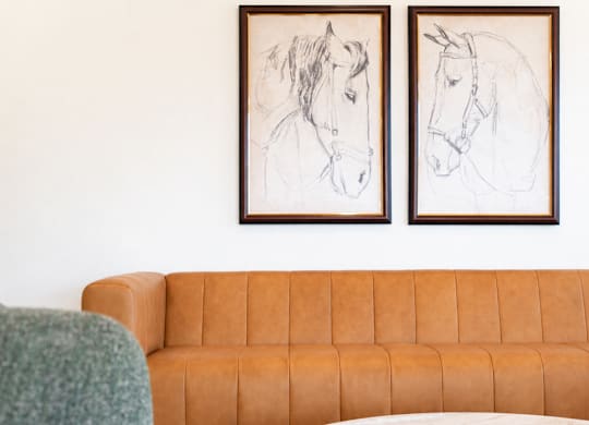 A living room with a brown couch, a green chair, and two framed pictures of horses on the wall.