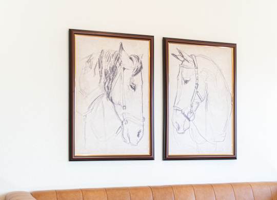 Two framed horse drawings hang on a wall above a tan couch.
