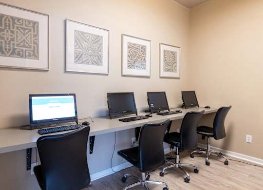 A row of computers are on a desk with three chairs.