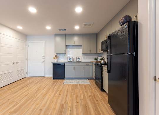 A black refrigerator is in the corner of a kitchen with wooden floors and white walls.