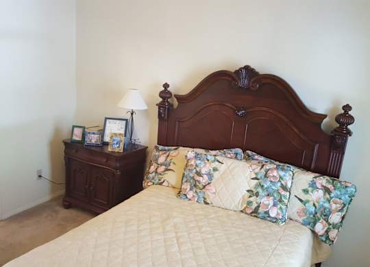 A bed with a floral pillow and a wooden headboard sits in a room with a nightstand and lamp.