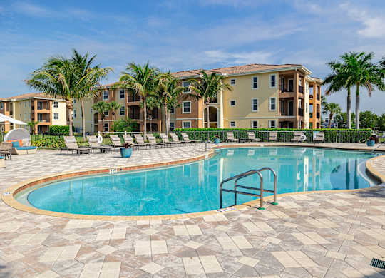 A swimming pool surrounded by a brick patio and palm trees in front of apartment buildings.
