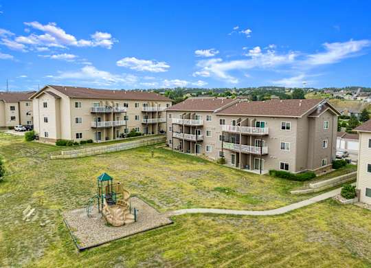 A playground with a swing set is in the middle of a grassy area in front of apartment buildings.
