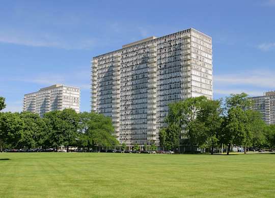 A large grassy field with trees and three buildings in the background.