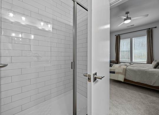 A white tiled bathroom with a white door and a bedroom with a bed and a ceiling fan.at Address East End, Raleigh North Carolina