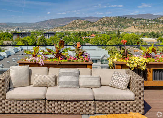 A wicker sofa with cushions is placed on a balcony with a view of the city and mountains.