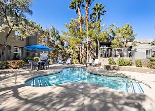 A pool surrounded by trees and chairs under a blue umbrella.