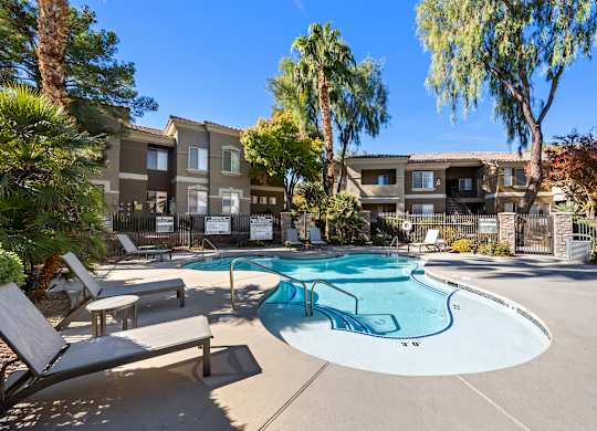 A swimming pool surrounded by palm trees and lounge chairs in front of apartment buildings.