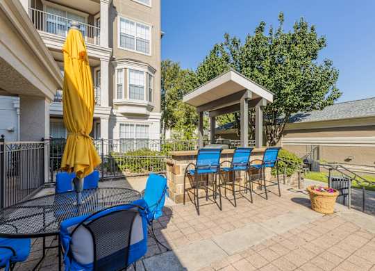 BBQ grill station seating area with blue stools and a table with chairs and closed yellow umbrella at Encore at Buckingham apartments in Richardson, TX