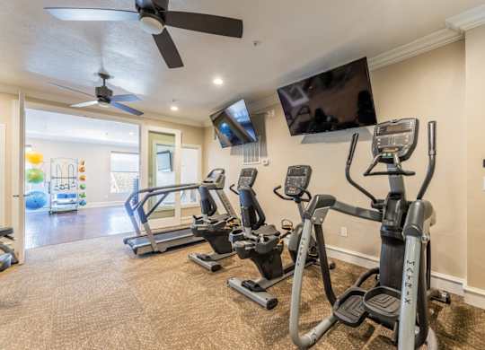 Cardio machines including an elliptical, cycling machines, and a treadmill facing a wall-mounted tv in the fitness center at Encore at Buckingham apartments in Richardson, TX