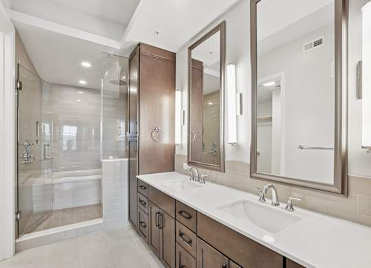 A modern bathroom from the C1 floorplan with a walk-in shower and tub, wood cabinets, sinks, and large mirrors at Alastair at Aria Village Apartments in Sandy Springs, GA