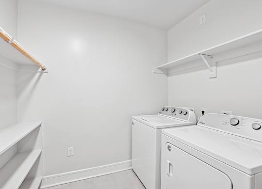 A white laundry room with a full washer and dryer and shelves at Alastair at Aria Village Apartments in Sandy Springs, GA