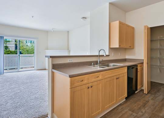 Soho kitchen with wooden cabinets, a pantry closet, and the living room in the background at Abbey Rowe Apartments in Olympia, WA