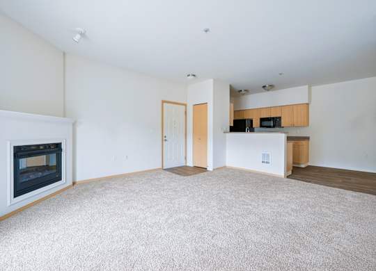 Spacious Soho living room with a fireplace, carpeting, and a kitchen in the background at Abbey Rowe Apartments in Olympia, WA