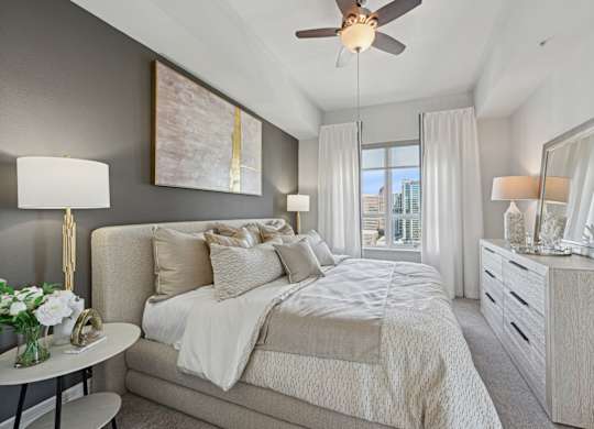 Model bedroom with a large bed, a ceiling fan, and a view of the city at Dominion Post Oak apartments in Houston, TX