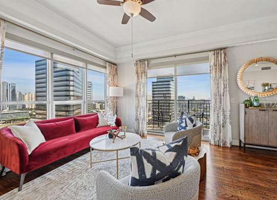Model living room with a red couch, a white coffee table, and a large window with a view of the city at Dominion Post Oak apartments in Houston, TX