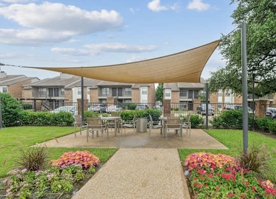 A shaded, outdoor dining area with lush grass and flowers surrounding it at Estelle Creek North Apartments in Irving, TX