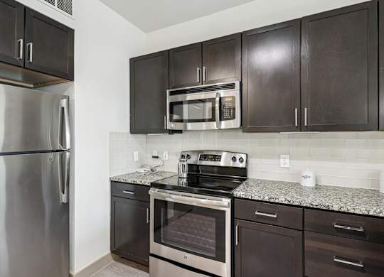 A kitchen with a stainless steel refrigerator, microwave, oven, and dark wood cabinets at GreenVue Apartments in Richardson, TX