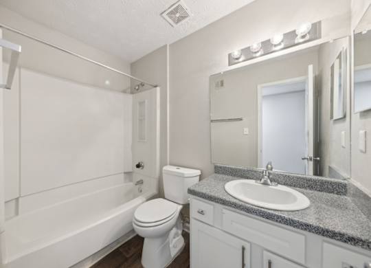 A bathroom with a white bathtub and shower, toilet, sink, and a mirror with lights at Gwinnett Square Apartments in Duluth, GA