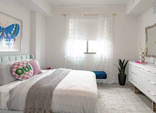 Side view of 1st bedroom in our 2-bed model with a white bed, a white dresser, and big window showering room with sunlight at Hollywood Heights on the Boulevard Luxury Apartments in Hollywood, FL