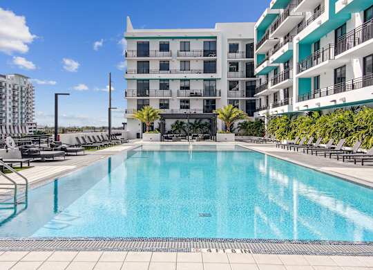 Large swimming pool surrounded by greenery and lounge chairs with a pergola in the background at Hollywood Heights on the Boulevard Luxury Apartments in Hollywood, FL