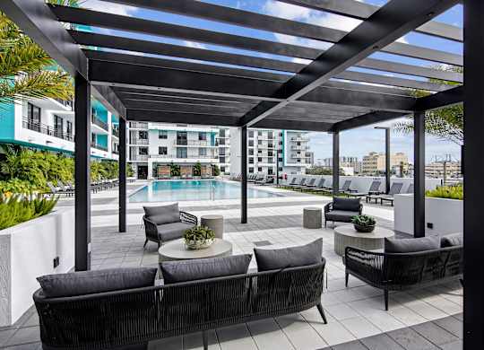 Outdoor couch seating under a pergola with our beautiful pool in the background at Hollywood Heights on the Boulevard Luxury Apartments in Hollywood, FL