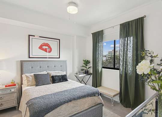 A bedroom with a large bed and a framed picture of red lips above it and a window with green curtains at Hollywood Heights on the Boulevard Luxury Apartments in Hollywood, FL