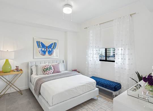 A bedroom with a bed, a blue bench, and a blue butterfly picture on the wall above the bed  Second bedroom with a bed, nightstand in our 2-bedroom model at Hollywood Heights on the Boulevard Luxury Apartments in Hollywood, FL