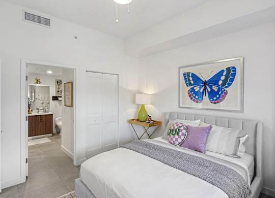 A bedroom with a bed, a blue bench, and a blue butterfly picture on the wall above the bed  Second bedroom with a bed, nightstand in our 2-bedroom model at Hollywood Heights on the Boulevard Luxury Apartments in Hollywood, FL