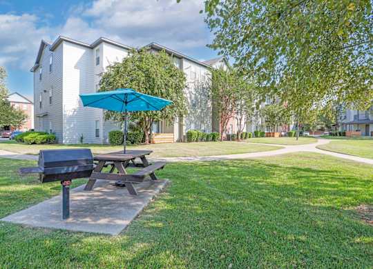 A picnic table with a blue umbrella is in the foreground of a grassy area with a building in the background.