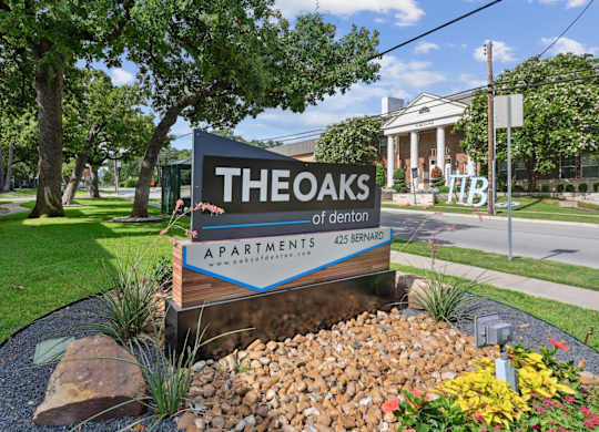 An entry sign for The Oaks of Denton Apartments is displayed in front of a landscaped area in Denton, TX