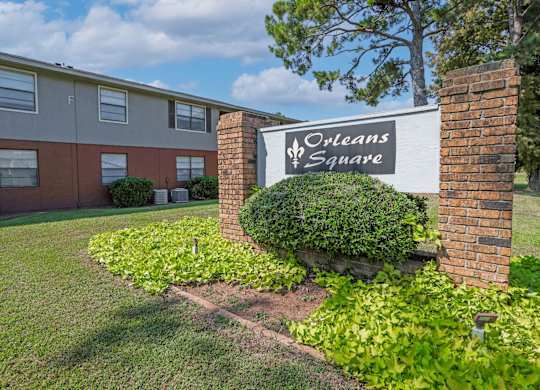 A sign for Orleans Square is surrounded by lush greenery in front of an apartment building at Orleans Square in Shreveport, LA