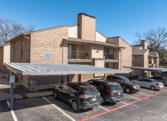 A covered parking area with cars and an apartment building in the background at Preston Oaks Apartments in Dallas, TX.