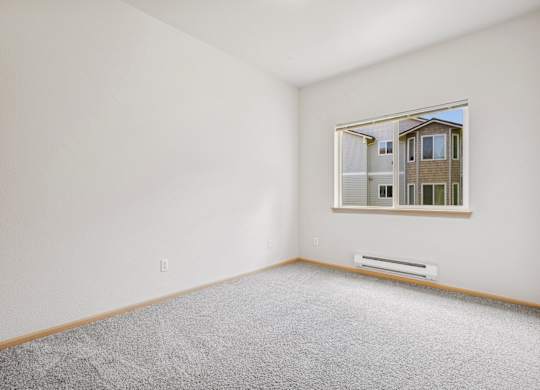 A bedroom with a carpeted floor and a window showing the apartment complex outside at The Madison Apartments in Olympia, WA