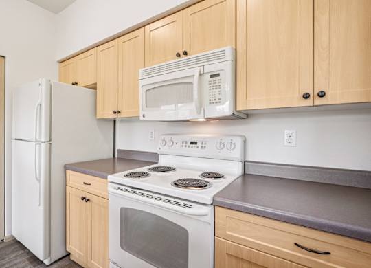 A kitchen with a white refrigerator, microwave, stovetop oven, and wooden cabinets at The Madison Apartments in Olympia, WA