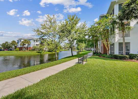 A walkway with a bench and a pond in front of apartment buildings at Wynnfield Lakes Apartments in Jacksonville, FL