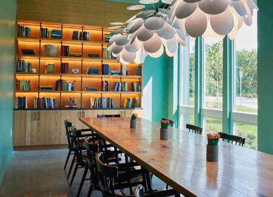 A long wooden table is surrounded by chairs in a room with a green wall and a bookshelf.