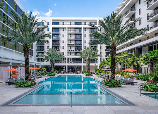 A large swimming pool surrounded by palm trees and lounge chairs in front of a multi-story building.