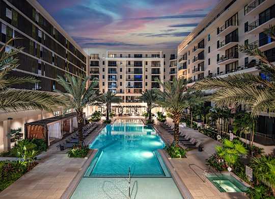 A large swimming pool surrounded by palm trees and lounge chairs in a hotel courtyard.