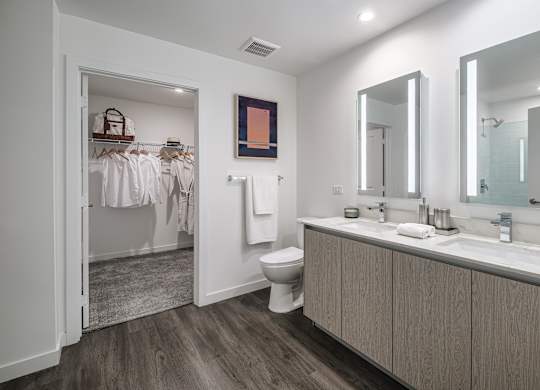 A bathroom with a toilet, sink, mirrors, and a closet with clothes hanging.