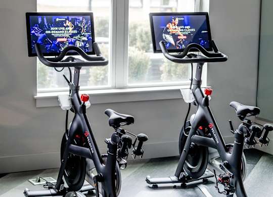 Two stationary bikes in a room with screens on the handlebars.
