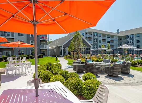 A bright orange umbrella is in the foreground of a sunny day at a courtyard with tables and chairs.