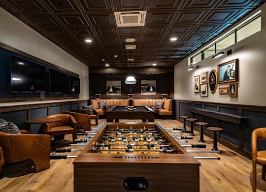 A foosball table is in the middle of a room with leather chairs and a bar area.