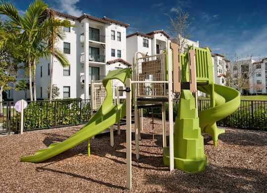 A playground with a green slide in front of apartment buildings.