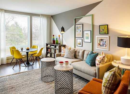 A living room with a grey couch, a black and white ottoman, a yellow chair, a black table, a bookshelf, and a window with white curtains.