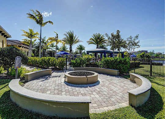 A circular brick patio with a fire pit in the middle, surrounded by a low wall and palm trees.